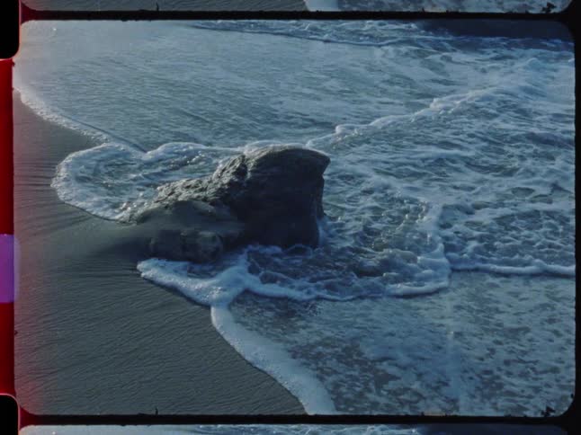 Vintage film of serene ocean waves washing over a rock on a sandy beach