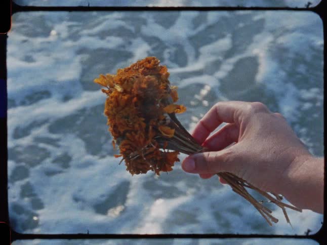 A hand holds a bouquet of wilting orange flowers over the ocean waves in a vintage film clip
