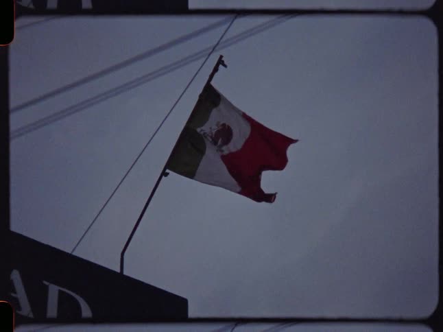 A vintage, grainy shot of the Mexican flag waving in the wind against a grey, overcast sky