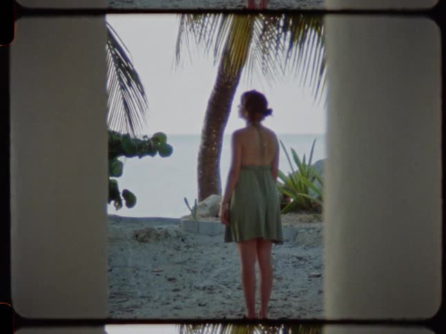 Vintage film footage frames a woman in a green dress from behind as she stands peacefully on a sandy beach looking out at the ocean