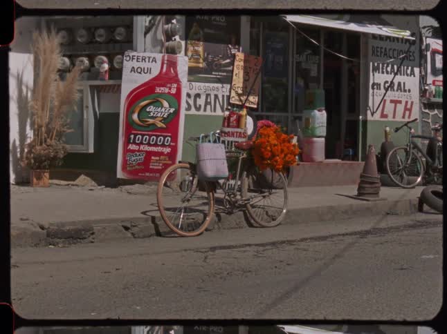 A vintage bicycle decorated with marigold flowers stands parked on a city street in front of a mechanic's shop as a car drives by