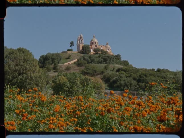 Vintage 16mm film scan of the Santuario de Nuestra Señora de los Remedios in Cholula, Mexico, with a field of cempasúchil flowers in the foreground