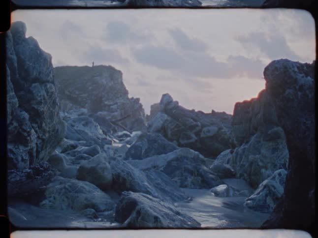 A dramatic, rocky coastline with a solitary figure on a distant cliff, captured on vintage Super 8 film at dusk
