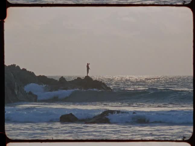 A lone fisherman casts a line from a rocky outcrop into the ocean as waves crash below in this vintage Super 8 footage