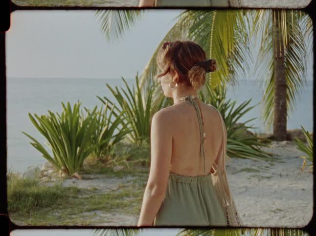 Back view of a woman in a summer dress standing on a sandy tropical beach looking out at the calm ocean