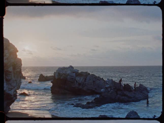 Super 8mm footage of people fishing on large rocks in the ocean as the sun sets