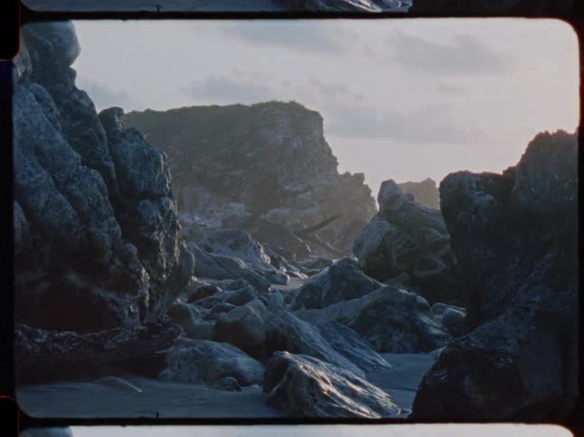 A vintage 8mm shot of a rugged, rocky coastline at sunset with gentle waves in the background