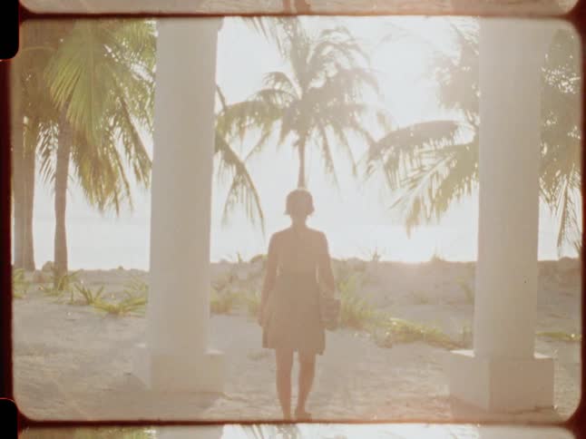 Vintage film shows a woman in a green sundress walking through white pillars toward a sunlit tropical beach