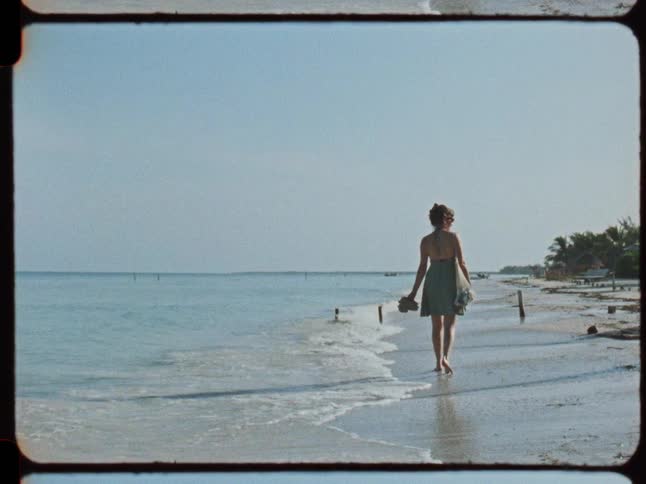 A vintage Super 8 film captures a woman in a summer dress walking barefoot along the shoreline of a serene tropical beach