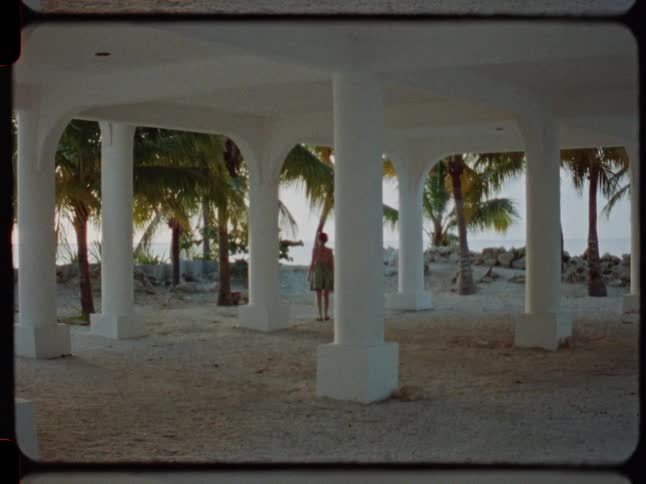 Vintage 8mm footage captures a woman walking away through a sandy, pillared ground floor of a beach house towards the ocean