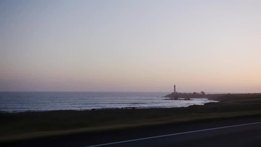 A scenic drive along a coastal highway reveals a distant lighthouse standing against the hazy sunset