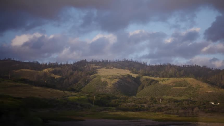 A scenic journey past rolling green hills under a dramatic cloudy sky, viewed from a moving window