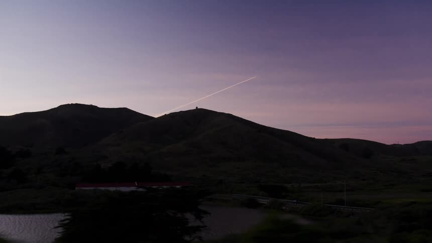 A jet contrail streaks across a beautiful purple twilight sky above silhouetted mountains, captured from a moving vehicle