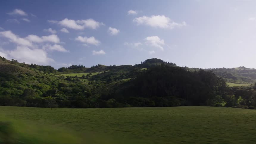 Point of view from a moving vehicle driving past a beautiful landscape of rolling green hills and lush forests under a blue sky