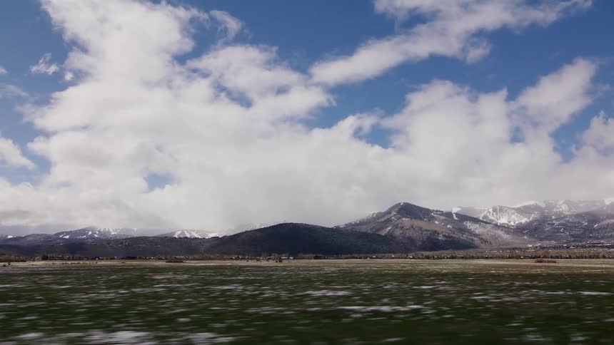 Scenic drive past a late-winter landscape featuring a vast field and snow-covered mountains under a partly cloudy sky