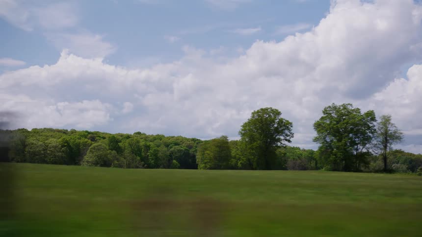 A fast-moving view from a vehicle window reveals a peaceful green countryside with fields and trees under a blue and cloudy sky
