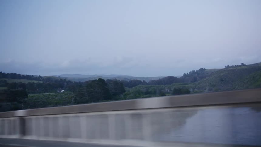 A moody point-of-view shot from a moving car captures the scenic rolling green hills and a winding river of the California coast on an overcast day