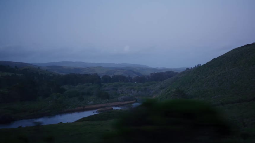 A slow-motion drive reveals a serene river winding through rolling green hills at dusk