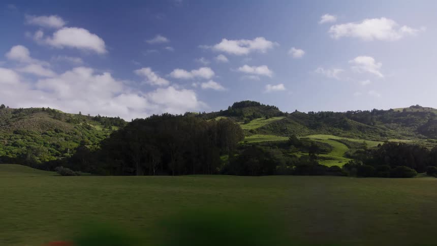 A point-of-view shot from a moving vehicle captures a scenic drive through a rural landscape with rolling green hills and a tranquil pond