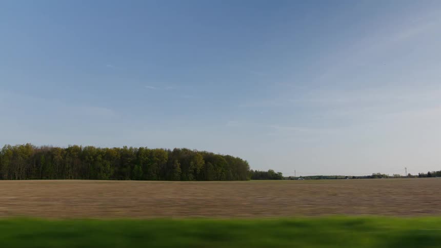 A picturesque view from a moving car window shows a vast, tilled farm field alongside a dense forest on a clear day