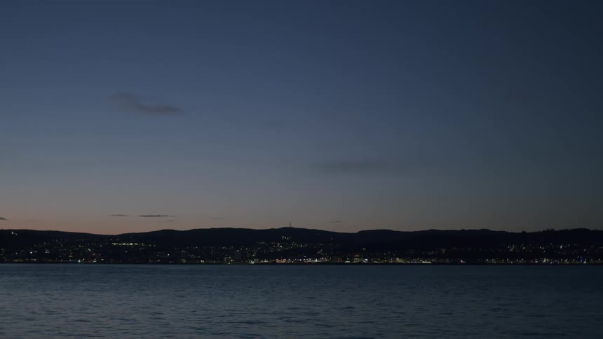 A serene wide shot of a coastal city glowing across the water during the blue hour