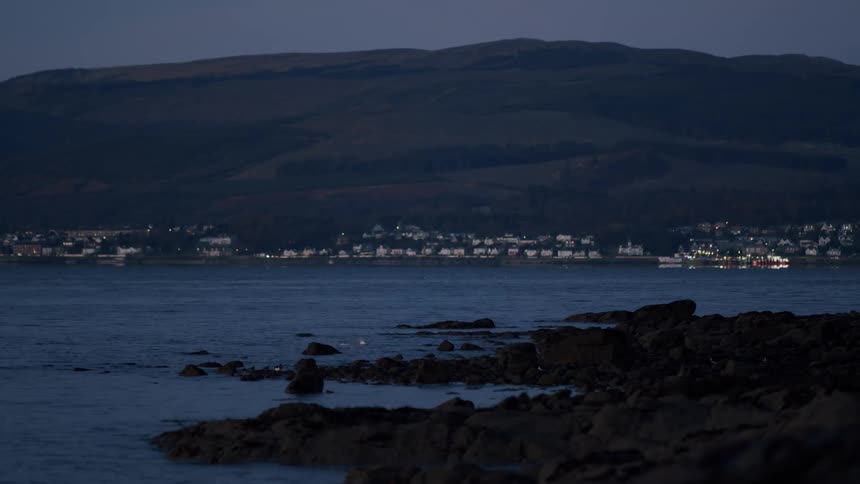 A quiet coastal town's lights glow across the bay from a rocky shoreline as dusk settles over the mountains