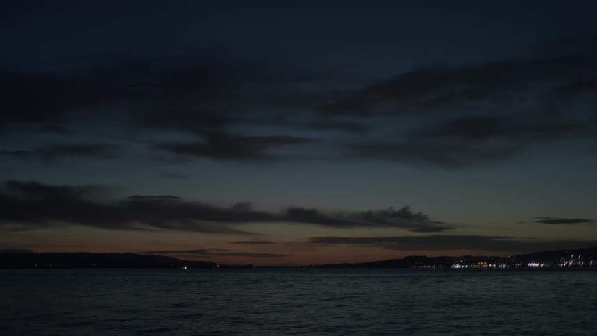 A tranquil sea at dusk with the last glow of sunset on the horizon and distant city lights along the coastline