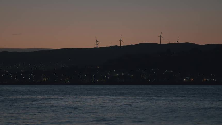 A tranquil dusk view across the water shows glowing city lights and silhouetted wind turbines spinning on the distant hills