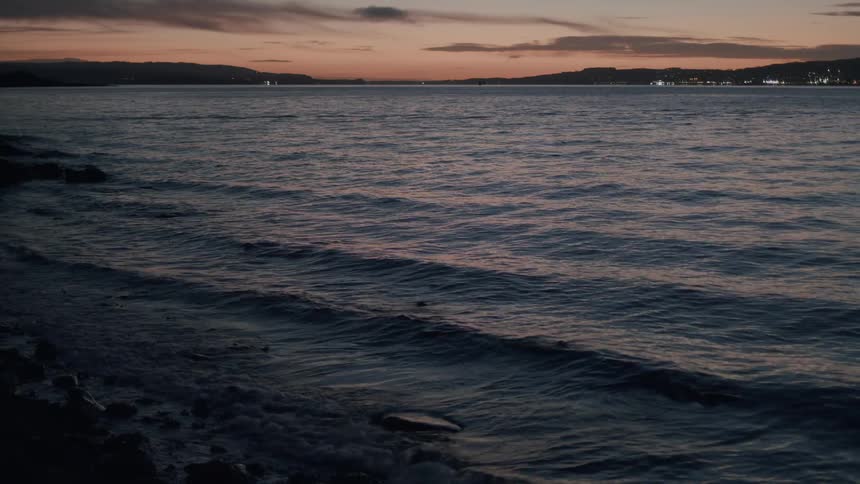 Calm ocean waves gently roll onto a rocky shoreline at twilight with distant city lights on the horizon
