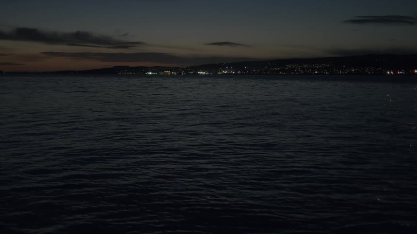 A calm sea at night with the glittering lights of a distant coastal city on the horizon