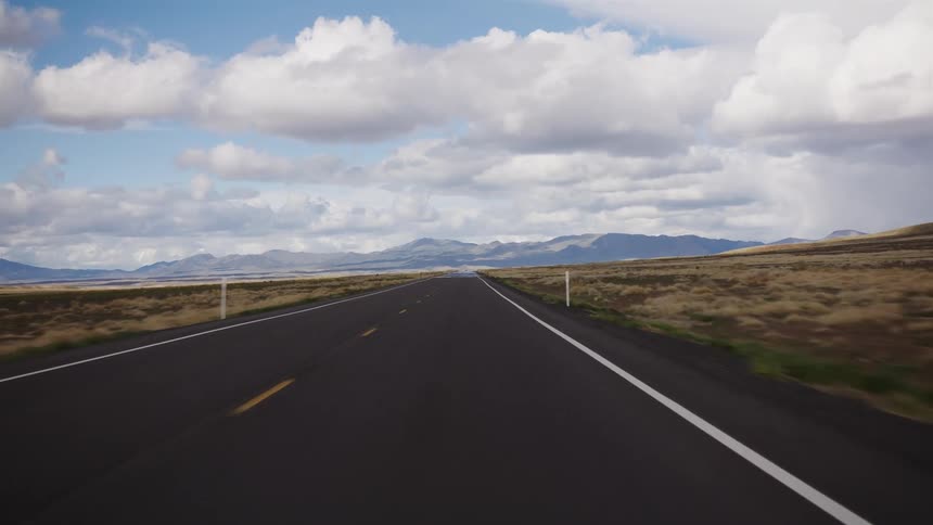 A point-of-view shot traveling down an empty, open road through a vast, rural landscape with mountains in the distance