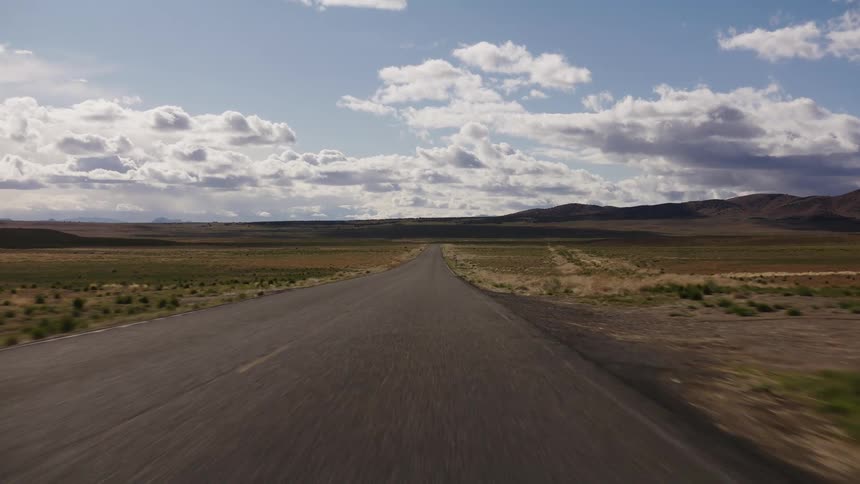 A point-of-view shot driving down a long, straight, and empty desert highway that stretches into the distant hills under a big, cloudy sky