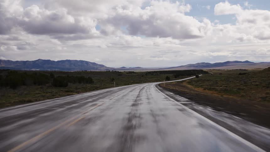 A point-of-view perspective of a vehicle driving quickly down a wet, winding rural highway through a vast, open landscape with distant mountains under a dramatic, cloudy sky