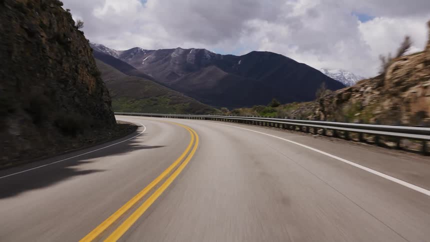 A point-of-view drive on a winding mountain road with majestic snow-capped peaks under a dramatic cloudy sky