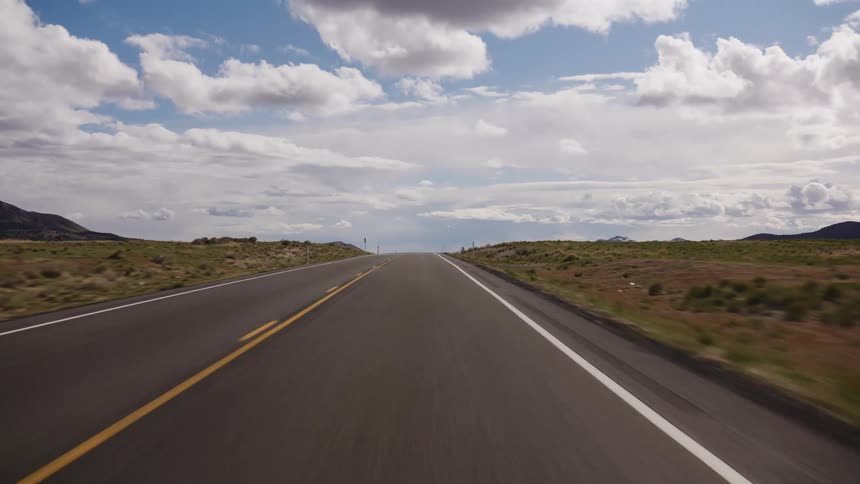 A point-of-view shot of driving along an endless, empty two-lane highway through a vast, open landscape under a cloudy blue sky