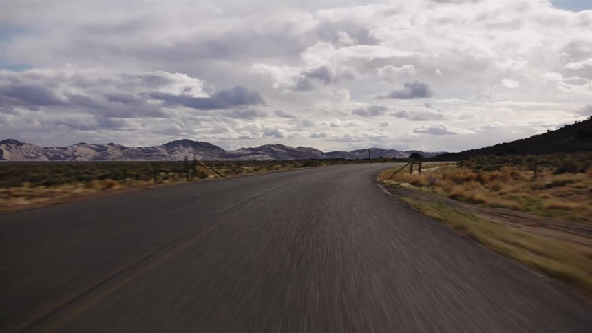 A fast-paced point-of-view drive down a winding, empty road through a vast desert landscape toward distant mountains under a dramatic, cloudy sky