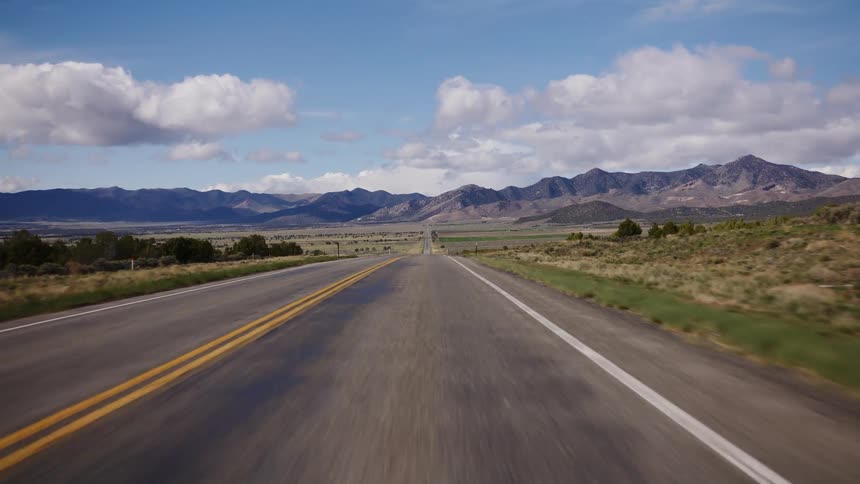 A scenic first-person view of driving down a long, straight desert highway towards distant mountains under a blue sky with fluffy white clouds