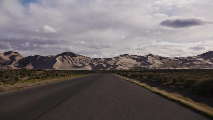 A low-angle, fast-moving perspective down a desolate desert road towards magnificent sand dunes