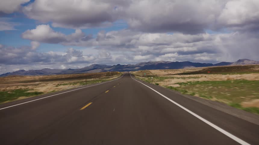 A first-person view of driving down a long, empty highway through a vast desert landscape towards distant mountains under a cloudy sky