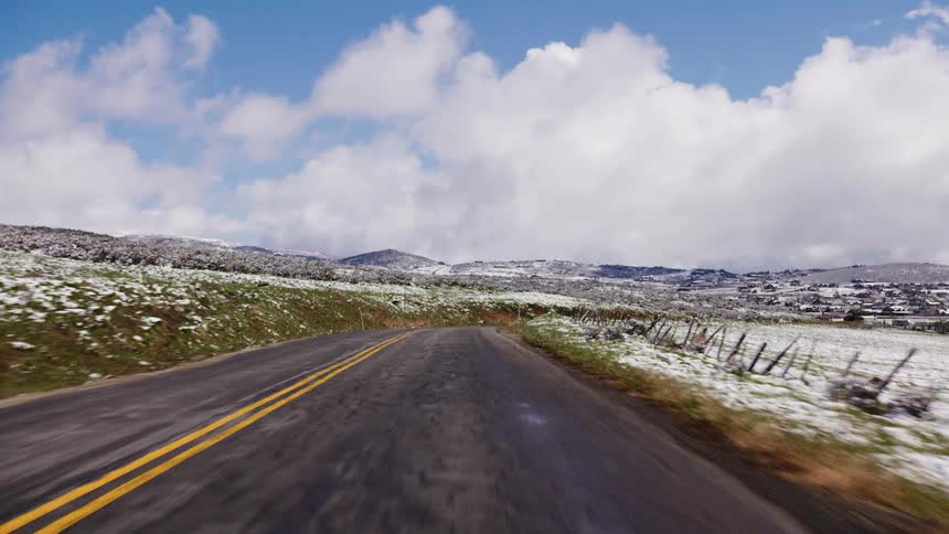 A scenic point-of-view drive on a country road through snow-dusted fields and hills toward a small town on a crisp, partly cloudy day