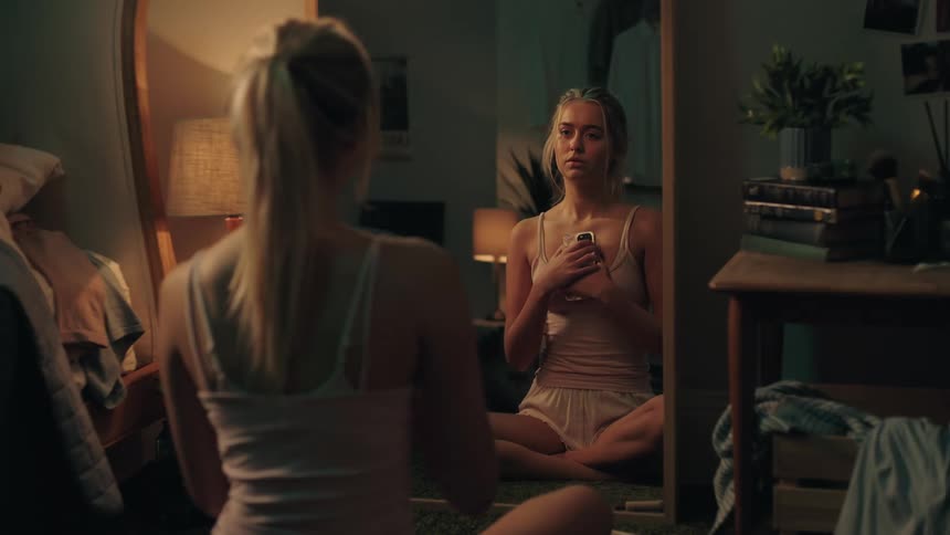 A distraught young woman sits alone in her dimly lit bedroom, holding her phone while looking at her sad reflection in the mirror