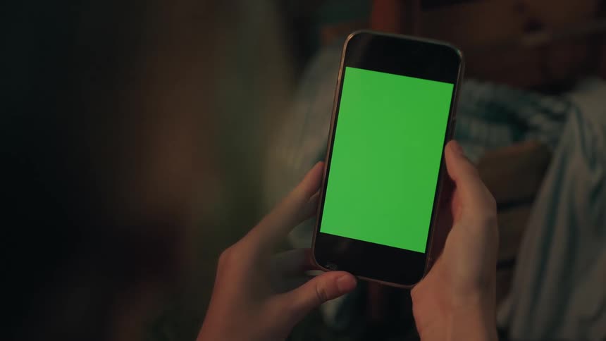 Over-the-shoulder shot of hands holding a smartphone with a vertical green screen in a dark room
