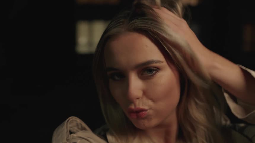 A playful blonde woman poses for the camera in a dimly lit setting, smiling, blowing a kiss, and making a peace sign in slow motion