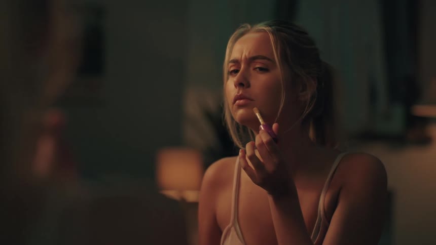 A young woman with a solemn expression looks in the mirror as she applies lip gloss in her dimly lit room