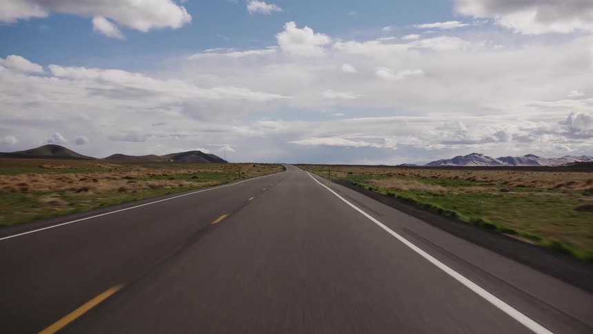 A driver's point-of-view traveling down a long, straight highway through a vast, open landscape with mountains in the distance