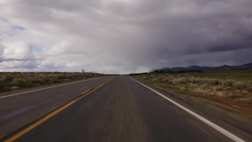 Point of view driving down an empty, open highway through the desert towards a looming storm in the distance