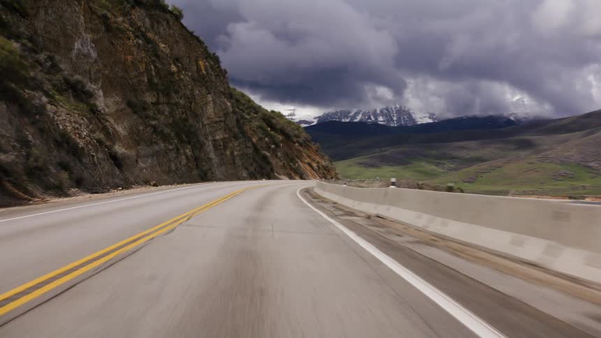 A scenic point-of-view drive along a winding mountain highway towards snow-capped peaks under dramatic, stormy clouds