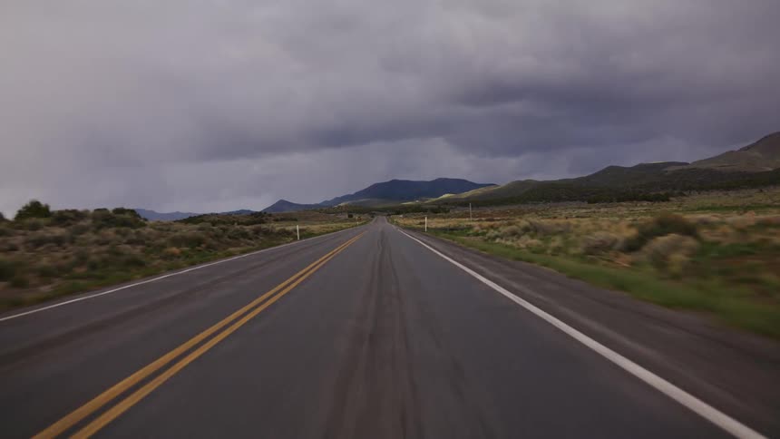 A point-of-view shot traveling down a remote, empty highway towards distant mountains under a dramatic, stormy sky