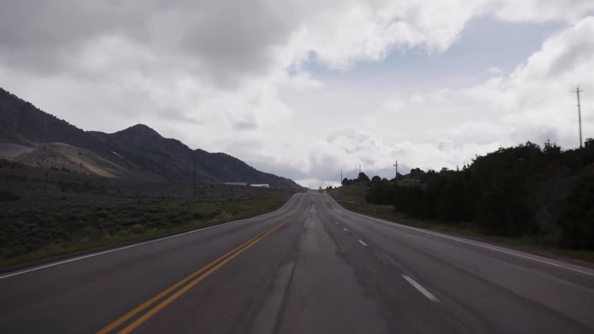 A POV shot driving down a long, empty highway through a mountainous and rural landscape under an overcast sky