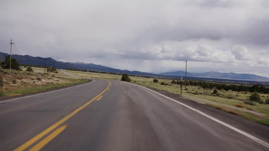 A point-of-view shot while driving down a rural highway through a vast, open landscape with mountains in the distance under a dramatic, cloudy sky